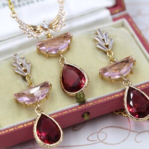 May include: A gold-toned jewelry set featuring a necklace and earrings, presented in a vintage pink jewelry box. The set includes a necklace with a pendant and earrings, each adorned with pink and red gemstones.