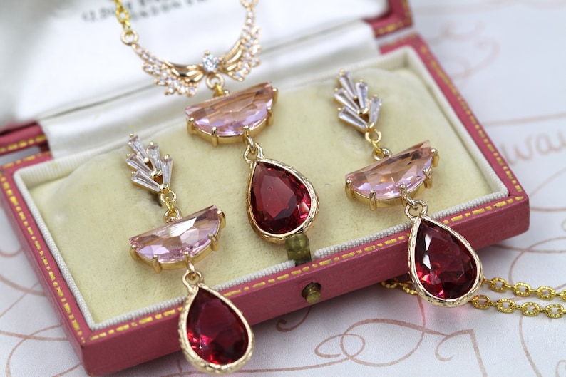 May include: A gold-toned jewelry set featuring a necklace and earrings, presented in a vintage pink jewelry box. The set includes a necklace with a pendant and earrings, each adorned with pink and red gemstones.