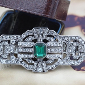 May include: A rectangular silver-toned brooch featuring a central emerald-green rectangular gemstone surrounded by numerous clear, sparkling rhinestones set in a geometric, symmetrical metal frame with intricate cut-out detailing and patterns.