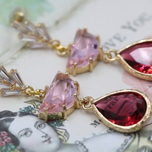 May include: A pair of gold-toned earrings with a stud backing. Each earring features a cluster of clear, rectangular crystals, a pink, triangular gem, and a red, teardrop-shaped gem. The earrings are set against a vintage-style background.
