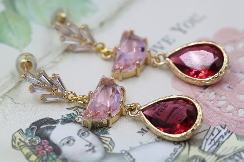 May include: A pair of gold-toned earrings with a stud backing. Each earring features a cluster of clear, rectangular crystals, a pink, triangular gem, and a red, teardrop-shaped gem. The earrings are set against a vintage-style background.