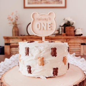 May include: A white frosted cake decorated with bear-shaped cookies. A wooden cake topper in the shape of a bear's head reads "Halfway to ONE". The cake sits on a wooden slab, perfect for a baby's half-birthday celebration.