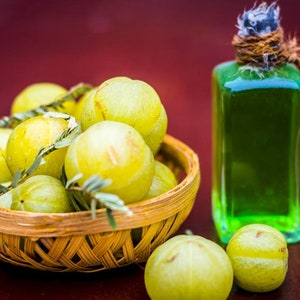 May include: A still life featuring a woven basket filled with yellow-green gooseberries and sprigs of greenery. A green liquid-filled bottle with a rustic twine-wrapped stopper sits nearby. The scene is set against a dark red background.