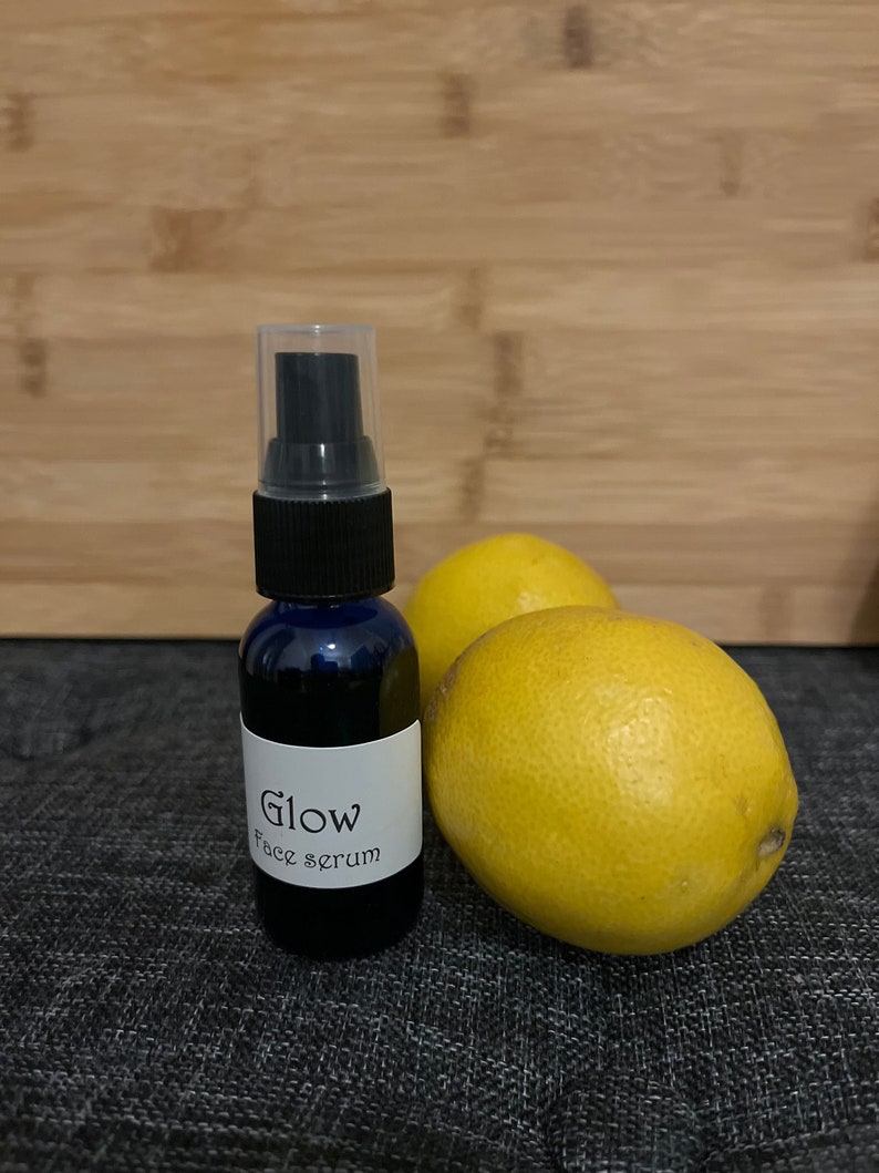 May include: A dark blue glass bottle with a black spray top and a white label that reads "Glow Face serum." Two bright yellow lemons are next to the bottle. The background is a wooden surface.