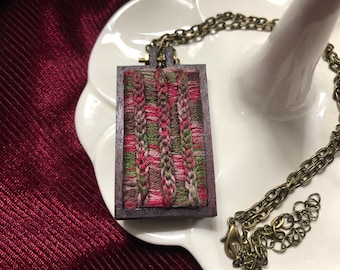 May include: A wooden framed pendant necklace with a colourful woven design. The pendant is attached to a brass chain.