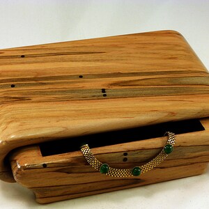 May include: A wooden jewelry box with a hinged lid and a curved front. The box is made of light wood with dark brown streaks and has a black velvet interior. A gold chain with green gemstones is inside the box.