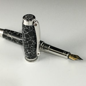 May include: A black and white marbled fountain pen with a silver cap and a gold nib. The pen is closed and lying on a white surface.