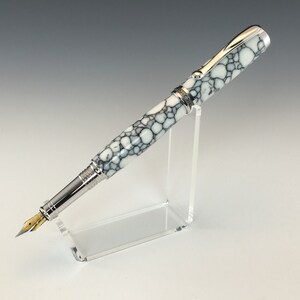 May include: A white and black marbled fountain pen with a silver trim and a gold nib. The pen is resting on a clear acrylic stand.