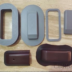 May include: A collection of grey and brown leathercraft components including a large grey oval frame, a grey rectangular block, a grey stitched border, a grey solid block, a brown rectangular leather piece with a hole, and a brown moulded leather case.