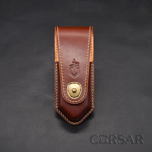 Pode incluir: Uma bainha de faca de couro marrom com fecho de pressão de latão. A bainha tem um design costurado e um logotipo em relevo na frente. O couro é de uma cor marrom escura rica.