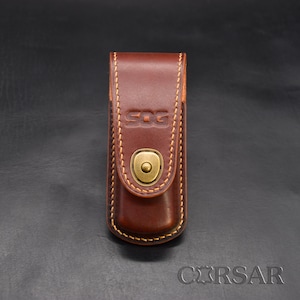 May include: A brown leather sheath with the embossed text "SOG" and a brass-colored snap closure. The sheath is stitched with light brown thread and appears to be designed to hold a knife or similar tool. The overall design is simple and functional.