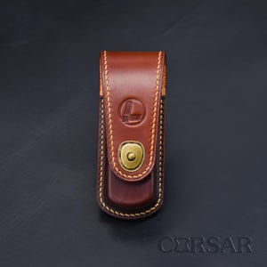 Peut inclure: Un étui en cuir marron avec une fermeture à rabat sécurisée par un bouton-pression en métal couleur laiton. Le cuir présente des coutures jaunes visibles sur les bords et un logo circulaire gaufré sur le rabat. Le mot CORSAR est inscrit.