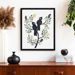 May include: A framed illustration of two black cockatoos perched on a branch with yellow flowers. The birds have yellow eyes and are surrounded by green leaves.