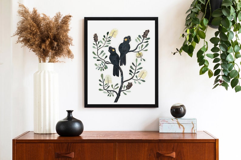 May include: A framed illustration of two black cockatoos perched on a branch with yellow flowers. The birds have yellow eyes and are surrounded by green leaves.