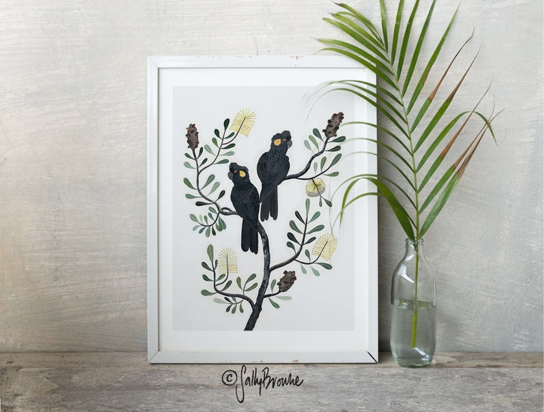 May include: A framed print of two black cockatoos perched on a branch of a tree with yellow flowers. The print is on a white background and has a white frame. The artist's signature is in the bottom right corner.