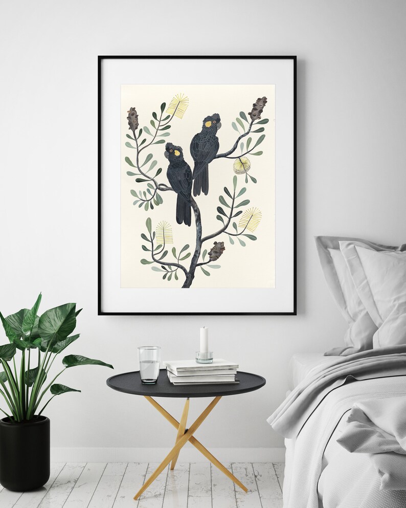 May include: A framed print of three black cockatoos perched on a branch of a stylized tree with yellow flowers. The background is a light cream color.