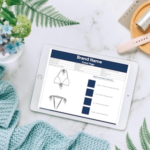 May include: A digital tablet displaying a swimwear design cover page with the brand name. The image includes a bikini top and bottom sketch, fabric swatches, and text. Accessories include a watch, perfume, and a wooden tray.