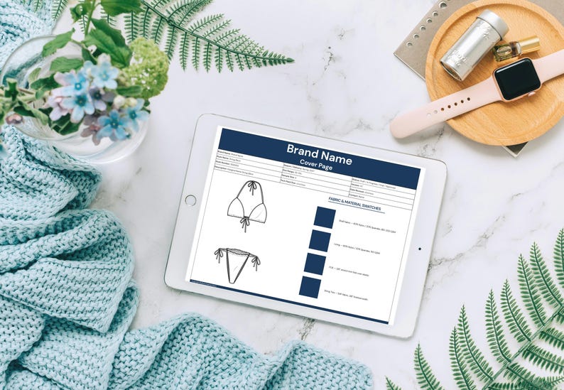 May include: A digital tablet displaying a swimwear design cover page with the brand name. The image includes a bikini top and bottom sketch, fabric swatches, and text. Accessories include a watch, perfume, and a wooden tray.