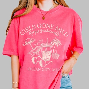 May include: A bright pink t-shirt with the text "GIRLS GONE MILD Rory's Bachelorette" and beach-themed graphics. The design includes a beach umbrella, lounge chair, sunglasses, a tropical drink, a palm tree, and a basket. The text "July 2025" and "OCEAN CITY, MD" are also printed on the shirt.