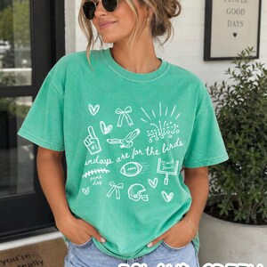 Pu&ograve; includere: Una t-shirt a maniche corte color verde isola con grafiche a linee bianche che includono un dito in gommapiuma, fiocchi, cuori, un pallone da football, un casco, una porta da gioco e il testo "Sundays are for the birds" con uno schema.