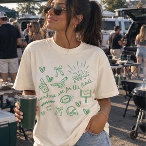 Pu&ograve; includere: Una t-shirt a maniche corte color crema con illustrazioni grafiche verdi a tema football americano, tra cui un casco, un pallone, un palo della porta, un dito di gommapiuma e fiocchi, con la scritta "sundays are for the birds" in verde.