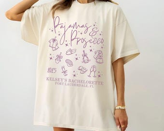 May include: A white oversized t-shirt with a purple graphic design that says "Pajamas & Prosecco" with a variety of small illustrations of pajamas, slippers, champagne glasses, and bows. The text "2023 Kelsey's Bachelorette Fort Lauderdale, FL" is also printed on the shirt.