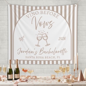 May include: A white and beige banner with the text "Vino Before Vows" and "Jordan's Bachelorette" is displayed. The banner includes the year 2026 and a graphic of two clinking champagne glasses. The banner is hanging above a table with champagne bottles and glasses.