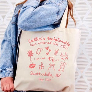 Peut inclure: Un sac fourre-tout beige avec un motif rose. Le texte dit "Caitlin's Bachelorette has entered the villa" avec des illustrations d'un palmier, d'un téléphone, de coeurs, d'une boisson, de lunettes de soleil, d'un bikini, de rouge à lèvres, d'un nœud et de coupes de champagne. L'emplacement et la date sont également imprimés.