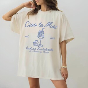 May include: Cream-colored t-shirt with the text "Ciao to Miss" in blue script. Below the text is a blue graphic of a cocktail in a glass with a lemon slice and straw. The text "Sophia's Bachelorette St. Petersburg, Florida" is also printed on the shirt.