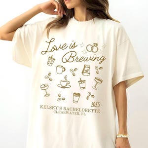 May include: Cream-colored t-shirt with the phrase "Love is Brewing" in a script font, along with coffee-themed illustrations. Includes the text "Kelsey's Bachelorette Clearwater, FL 2025". The design features coffee cups, beans, and champagne glasses.