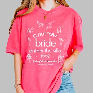 May include: A hot pink t-shirt with white text and illustrations. The text reads "a hot new bride enters the villa" with additional text below indicating "kelsey's bachelorette ocean city, md". The shirt features drawings of a phone, sunglasses, and other beach-themed icons.