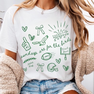 Pu&ograve; includere: Una t-shirt bianca a maniche corte con grafiche a linee verdi di icone a tema football americano, tra cui un dito di gommapiuma, un'aquila, un pallone, un casco, una porta da gioco e dei fiocchi. Il testo recita: sundays are for the birds, game day e Touch Down.