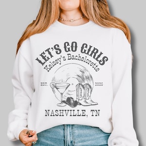 May include: White sweatshirt with the text "Let's Go Girls Kelsey's Bachelorette Est. 2025 Nashville, TN" printed on the front. The design includes a record, cowboy boots, a cowboy hat, and a cocktail glass.