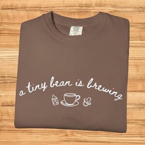 Może przedstawiać: Brązowa koszulka z białym napisem "a tiny bear is brewing". Projekt zawiera butelkę dla dziecka, filiżankę do herbaty i smoczek. Koszulka jest wykonana z miękkiego materiału.
