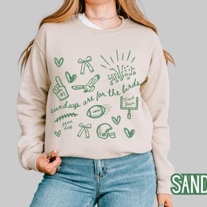 Pu&ograve; includere: Una felpa girocollo color sabbia con una stampa grafica verde scuro di disegni a tema football americano, tra cui un dito di gommapiuma, un pallone, un casco, una porta da gioco e il testo "sundays are for the birds" in corsivo.