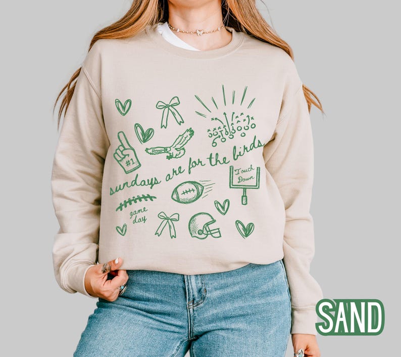 Pu&ograve; includere: Una felpa girocollo color sabbia con una stampa grafica verde scuro di disegni a tema football americano, tra cui un dito di gommapiuma, un pallone, un casco, una porta da gioco e il testo "sundays are for the birds" in corsivo.