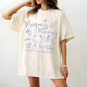 Puede incluir: Una camiseta blanca con un diseño gráfico azul que dice "Margaritas & Matrimony 2025" con varios iconos azules que incluyen una copa de margarita, un anillo, un zapato y una botella. El texto "Kelsey's Bachelorette" también está incluido en el diseño.