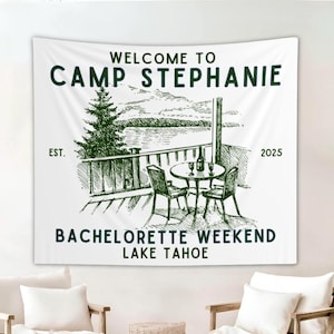 May include: White fabric wall hanging with a green illustration of a lakeside scene, including a table, chairs, and a pine tree. Text reads "WELCOME TO CAMP STEPHANIE BACHELORETTE WEEKEND LAKE TAHOE EST. 2025."