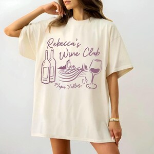 May include: A cream-coloured t-shirt featuring a purple graphic with the text "Rebecca's Wine Club Napa Valley, CA". The design includes wine bottles, a wine glass, and a vineyard. The shirt has short sleeves and a loose fit.