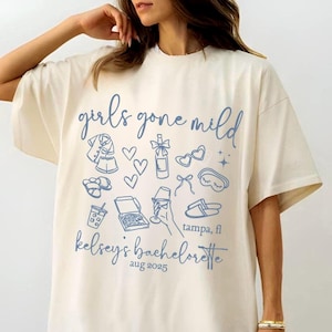 May include: Cream-colored t-shirt with the text "girls gone mild" in a script font. The shirt features light blue illustrations of pajamas, hearts, champagne, sunglasses, and other party-themed icons. Below, it reads "kelsey's bachelorette" and "aug 2025".
