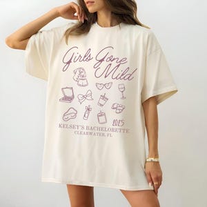 Puede incluir: Una camiseta blanca con un gráfico rosa que dice "Girls Gone Mild" e incluye varios iconos como gafas de sol, una copa de vino, un lazo y una vela. El texto "Kelsey's Bachelorette 2025 Clearwater, FL" también está impreso en la camiseta.