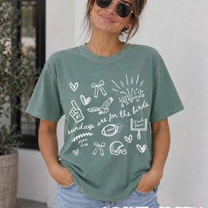 Pu&ograve; includere: Una t-shirt a maniche corte verde chiaro con grafiche a linee bianche che includono un pallone da football, un casco, una porta, un'aquila, fiocchi, cuori e il testo "sundays are for the birds" insieme a "game day" e "touch down" in corsivo.