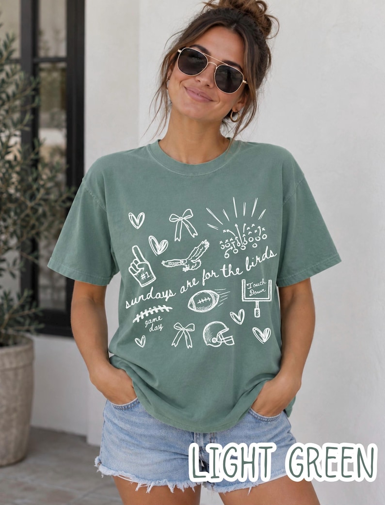 Pu&ograve; includere: Una t-shirt a maniche corte verde chiaro con grafiche a linee bianche che includono un pallone da football, un casco, una porta, un'aquila, fiocchi, cuori e il testo "sundays are for the birds" insieme a "game day" e "touch down" in corsivo.