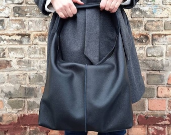 May include: A black leather tote bag with a long strap. The bag is being held by a person wearing a gray coat and blue jeans.