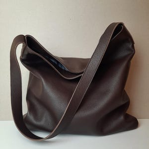 May include: A dark brown leather shoulder bag with a long strap. The bag has a soft, slouchy design and a textured surface. The strap is attached to the top of the bag and is designed to be worn over the shoulder.