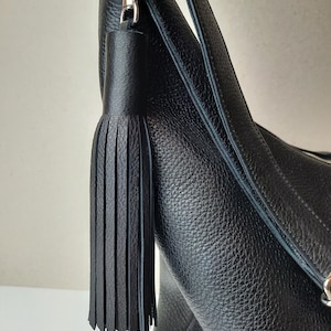 Könnte beinhalten: Nahaufnahme einer schwarzen Lederhandtasche mit silberfarbenem Verschluss und einer langen, schwarzen Quaste. Die Tasche hat eine strukturierte Oberfläche und einen Schulterriemen. Die Quaste besteht aus mehreren Lederstreifen.