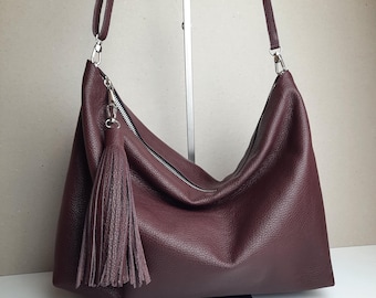 May include: A burgundy leather shoulder bag with a long strap and a silver zipper. The bag features a large tassel and a spacious design. The bag is made of a textured leather material.