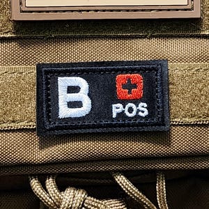 May include: Tan tactical backpack with two patches. The top patch is an American flag. The bottom patch is black with the letters "B POS" and a red cross symbol.