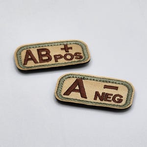 May include: Two rectangular patches with rounded corners. One patch reads "AB POS" and the other reads "A NEG". The patches have a tan background with brown text and a green border. The patches are likely for medical or tactical use.