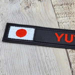Könnte beinhalten: Ein schwarzer, bestickter Aufnäher mit der japanischen Flagge und dem Namen "YUTO" in Rot. Der Aufnäher hat eine rechteckige Form und liegt auf einer Holzoberfläche.